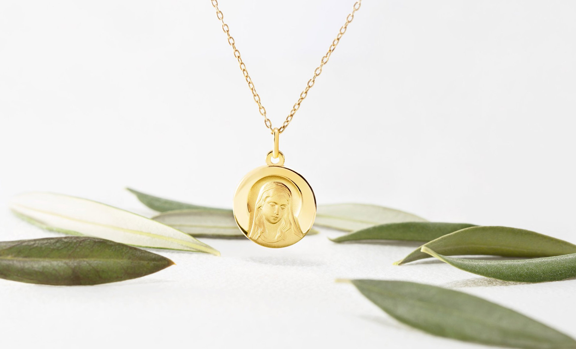 Medalla de virgen en oro con cadena recomendada como regalo de comunion, el colgante está sobre fondo blanco con hojas de color verde